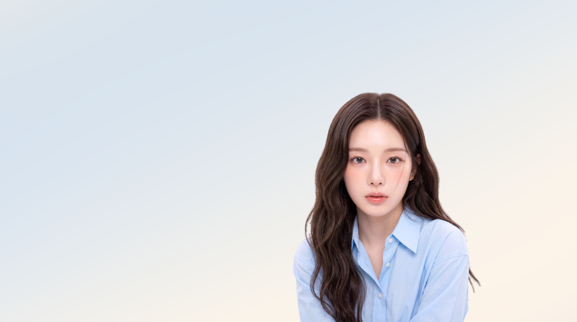 파우더블루에서 아이보리로 번지는 배경 앞에서 하늘색 셔츠를 입고 정면을 바라보는 긴 웨이브 머리의 여성 모델