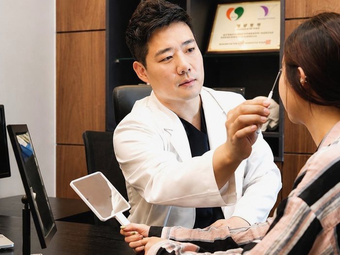 Bác sĩ chuyên khoa phẫu thuật thẩm mỹ cầm gương tay, ngồi đối diện bệnh nhân trong phòng khám và chỉ vị trí trên khuôn mặt để tư vấn